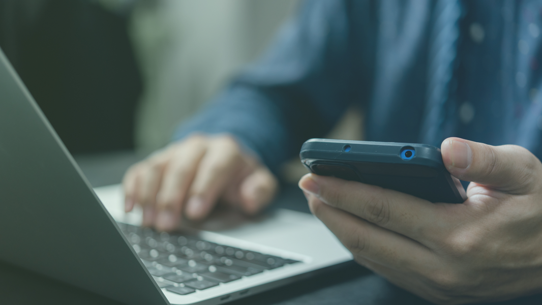 Person bedient Laptop und hält Handy in der Hand