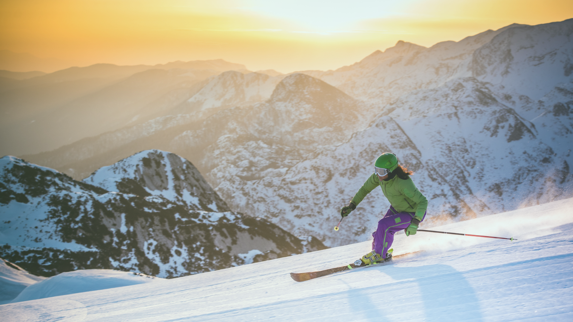 Skifahrer im grünen Anzug fährt eine verschneite Piste hinunter, dahinter ein sonniges Bergpanorama.