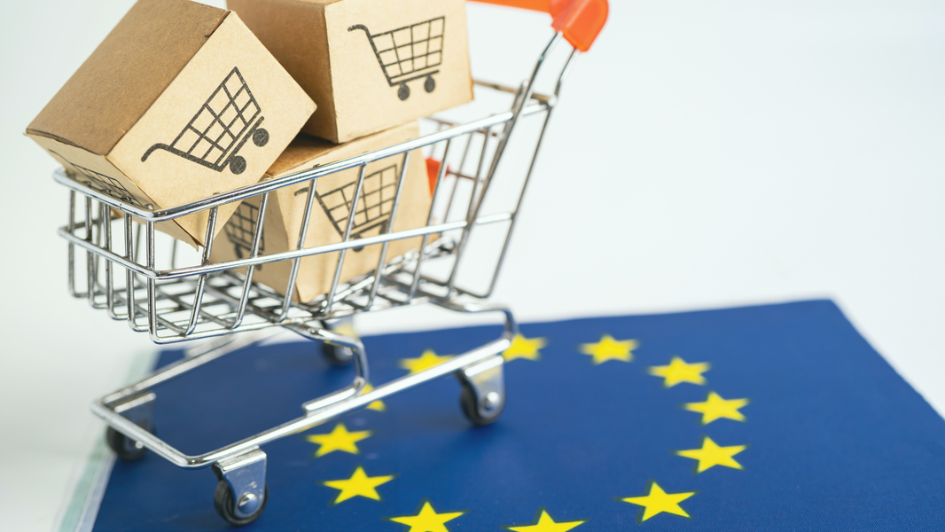 Ein Mini-Einkaufswagen aus Metall mit Paketen, auf denen Einkaufswagen-Symbole zu sehen sind, steht auf einer EU-Flagge. Das Motiv steht für Onlinehandel und Produktsicherheit in der EU.